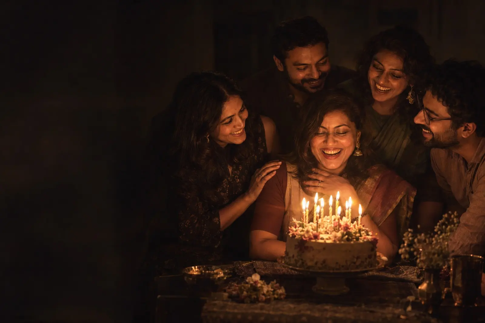 Made-up friends gathered close around a woman smiling at her candlelit birthday cake